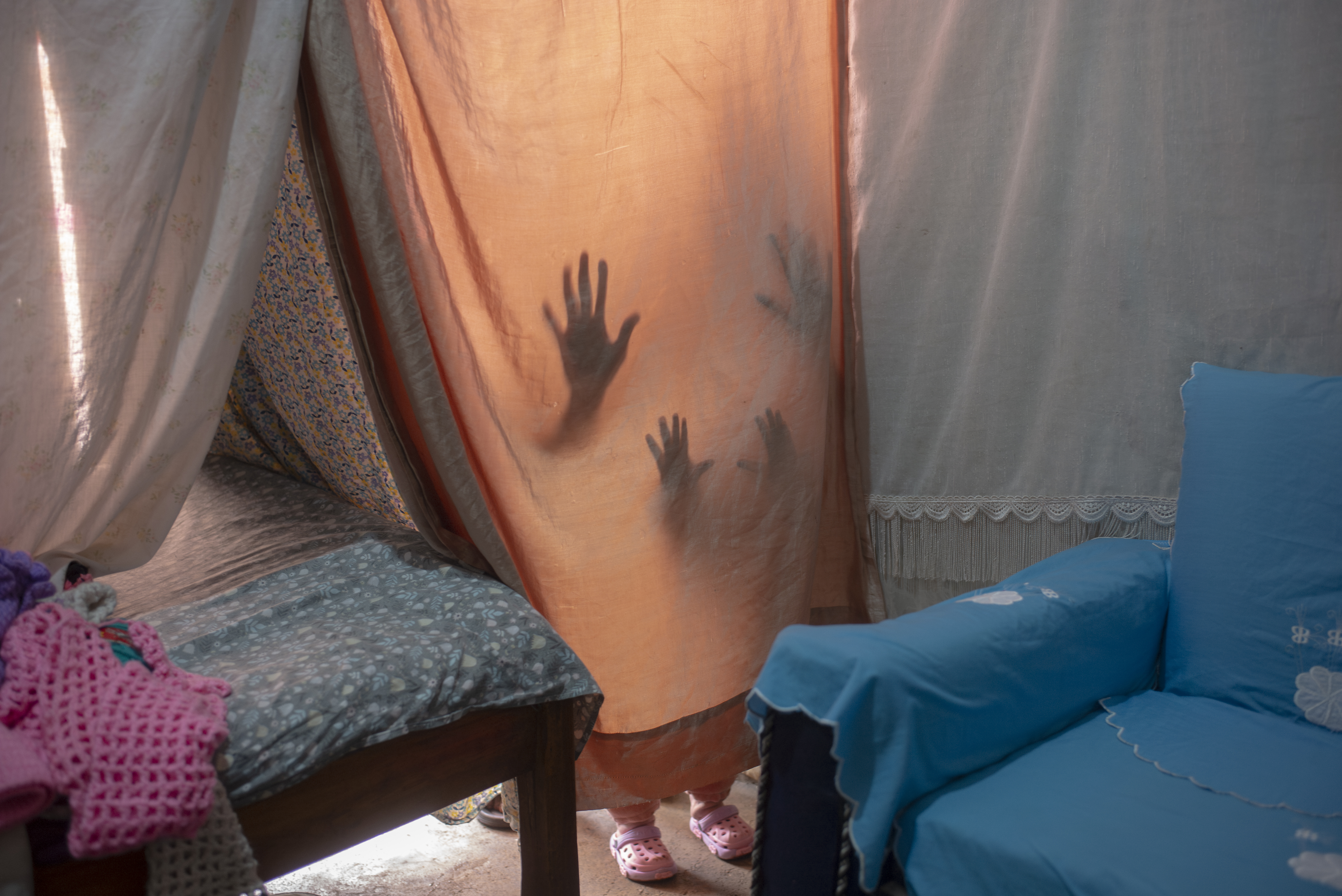 two pairs of hands (an adult's and a young child's) cast as silhouettes behind a curtain blocking the entrance to a humble room 