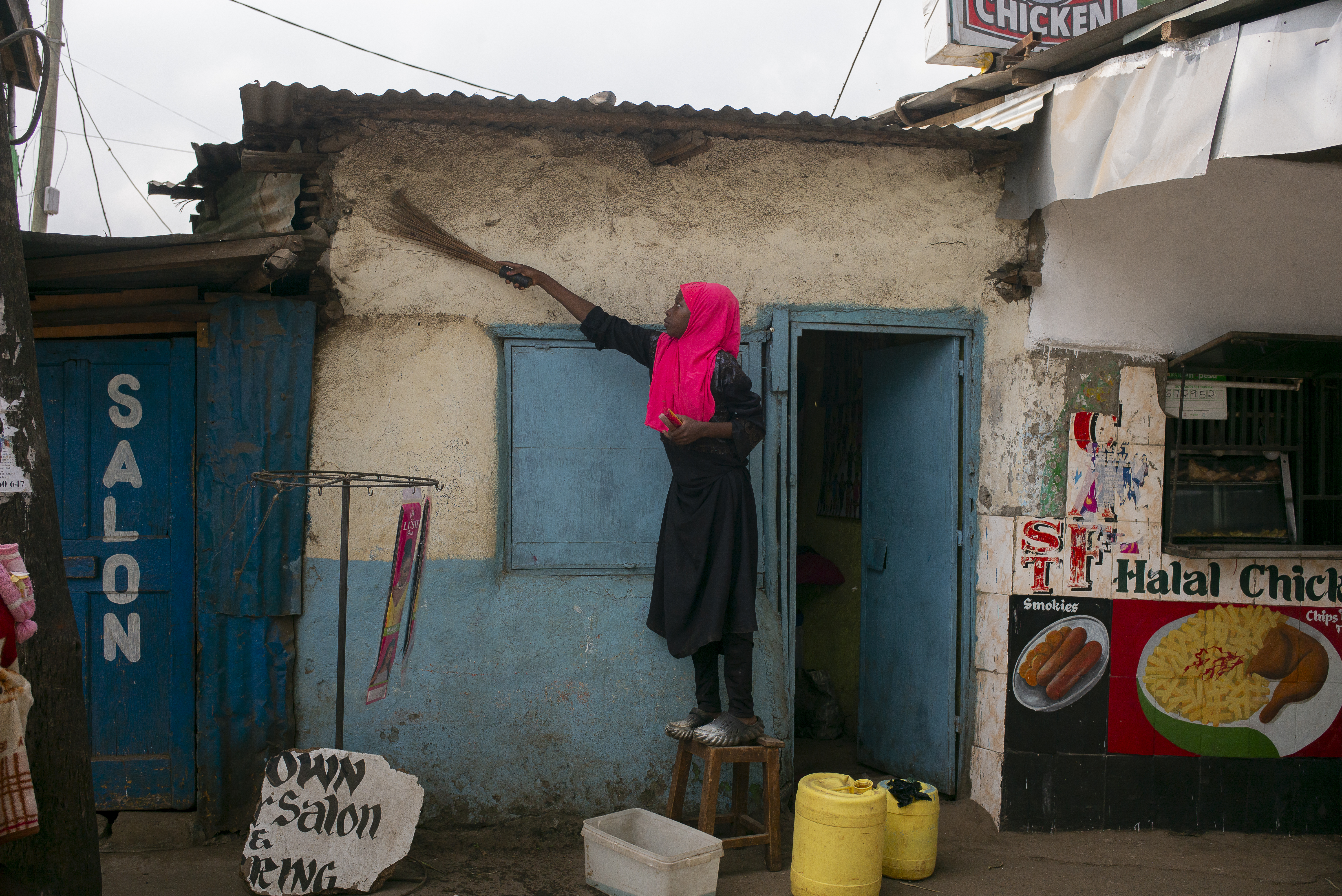 A young girl standing on a stool, wearing a pink scarf on her head, is cleaning the front of a store with a feather 