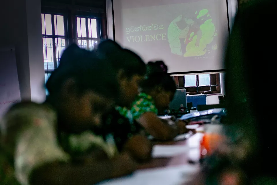 Participants silhouetted against bright windows listen as a facilitator stands at the front, the word "VIOLENCE" projected on the screen behind her.