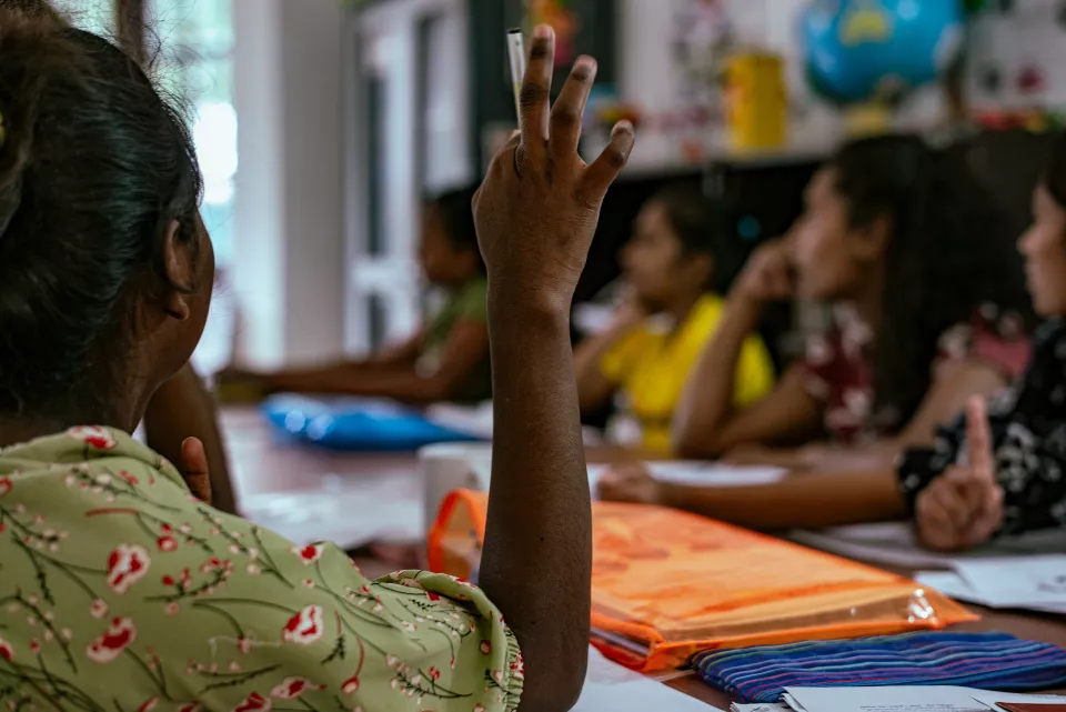 A woman raises her hand with a pen during a community meeting, other participants visible and attentive in the background.