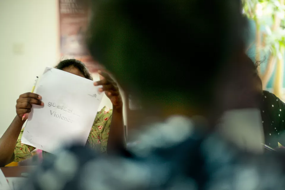 A woman holds up a sheet of paper bearing the word "Violence" in both Sinhala and English, her face hidden behind it &mdash; a striking image from what appears to be an awareness workshop.
