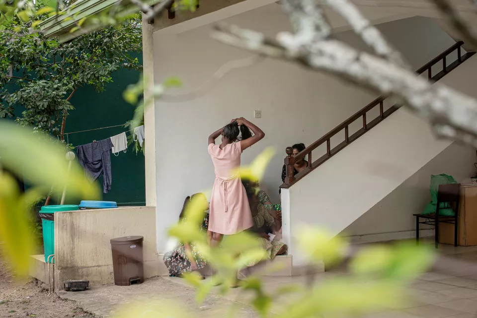 Framed through soft foliage, a woman in a pink dress ties her hair by a staircase while others sit nearby &mdash; a quiet, candid moment of everyday life in a shared courtyard.