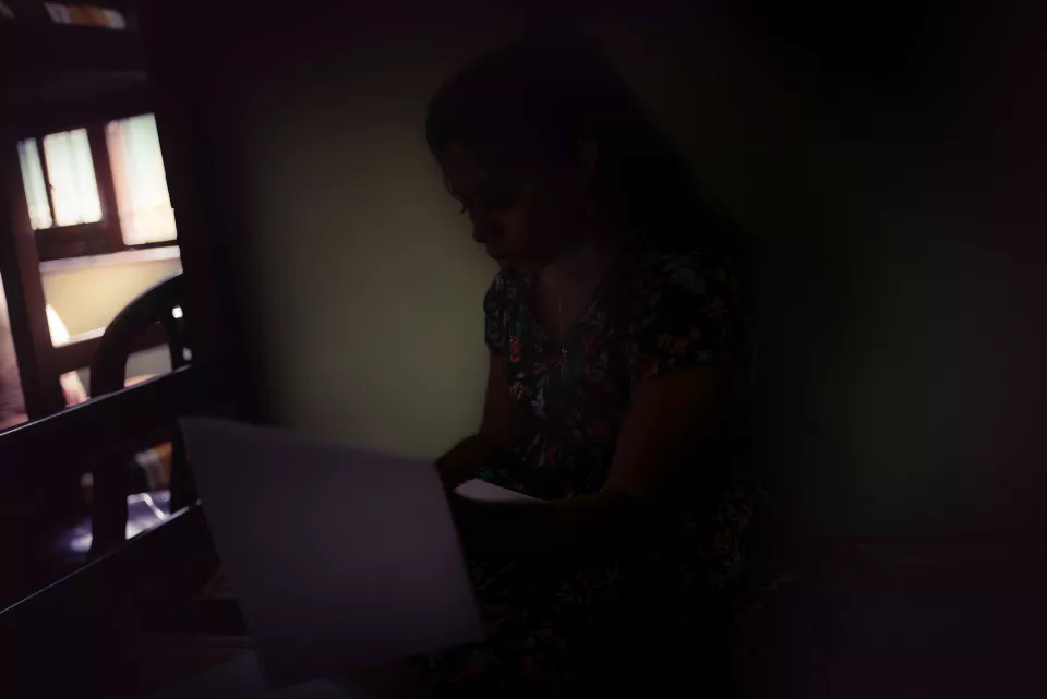 A woman in a floral dress reads papers alone in a darkened room, lit only by a faint glow from the window beside her.