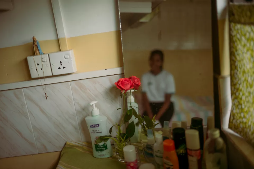 A bedroom vanity with red roses, toiletries, and a Virgin Mary figurine, a young woman's blurred reflection visible in the mirror behind.