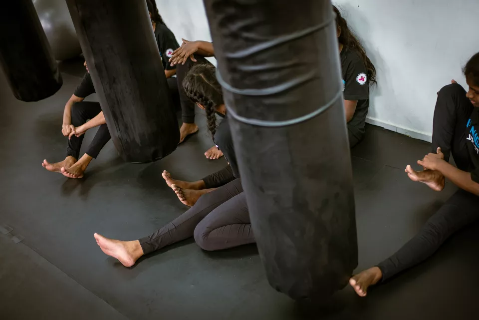 A group of girls in matching black uniforms rest between rounds on the mat of a boxing gym, heavy punching bags towering around them.