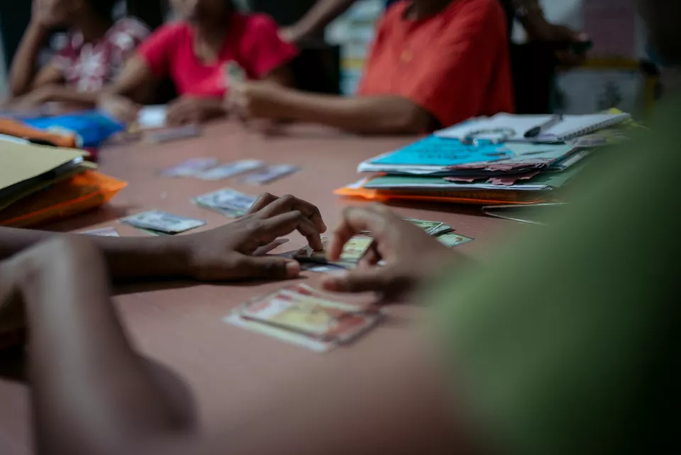 Hands sort through small cards and banknotes spread across a table during what appears to be a community savings or microfinance meeting, notebooks and folders stacked nearby.