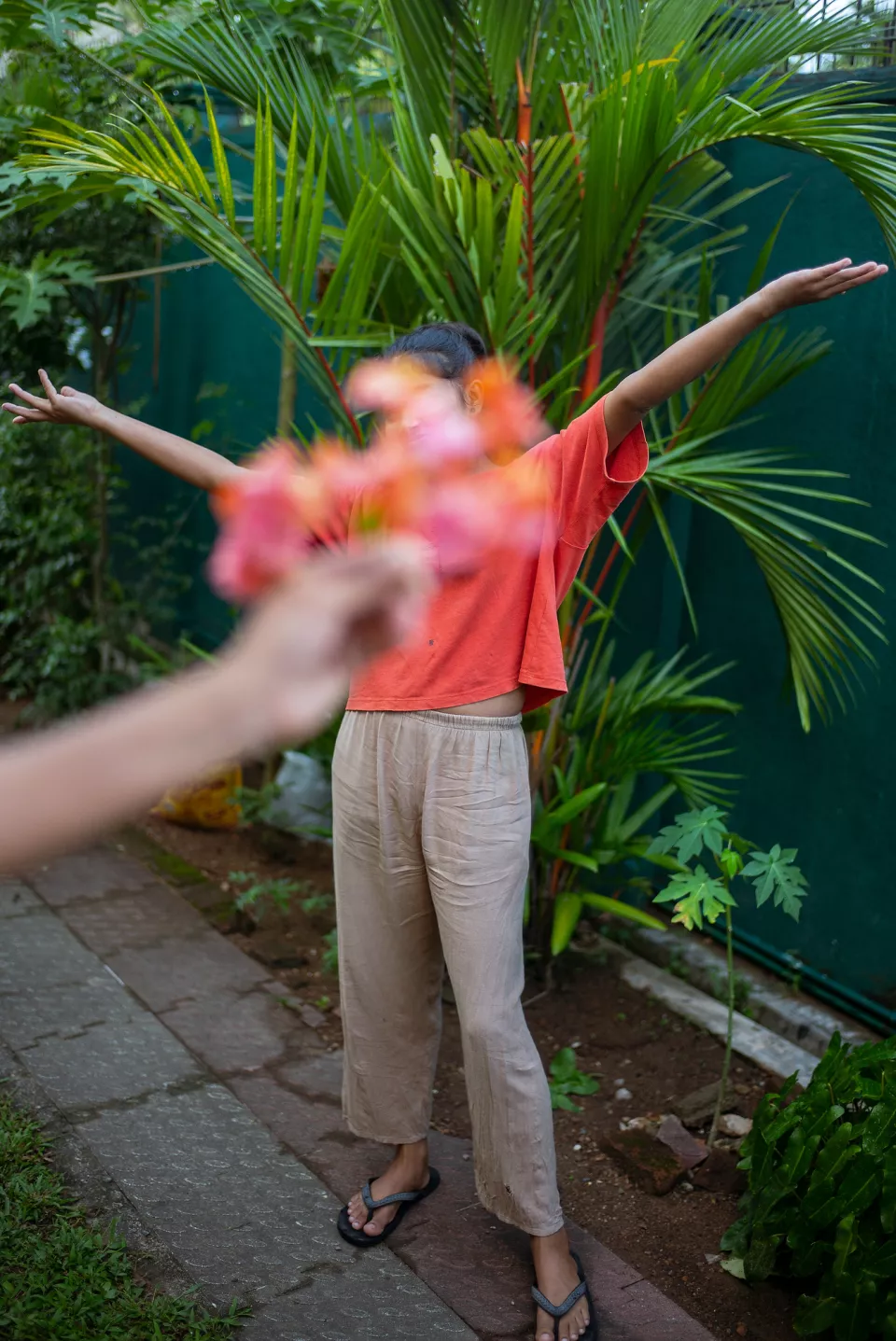 A young woman stretches her arms wide in a lush garden, her face hidden by a bouquet of tropical flowers held up by an unseen hand in the foreground.