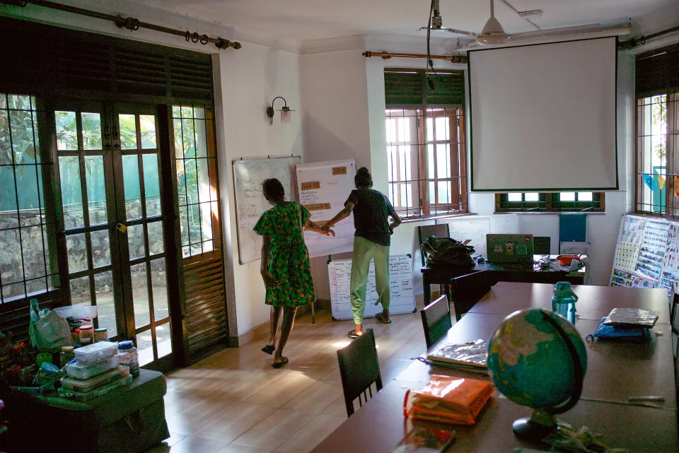 Two people collaborate at a whiteboard in a bright, airy colonial-style classroom, a globe and scattered books on the table in the foreground.
