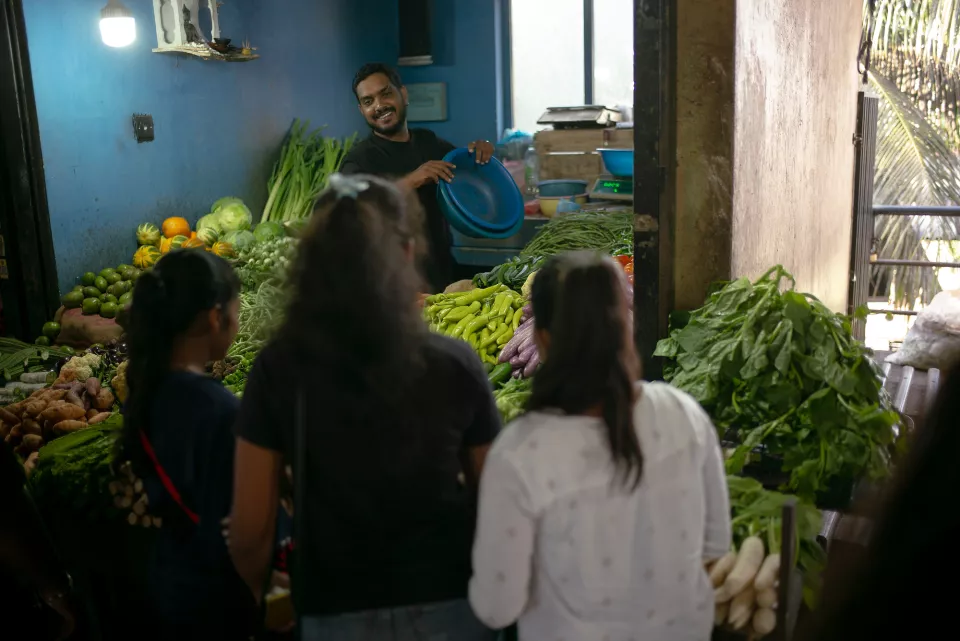 A smiling vendor holds up a blue bowl behind his stall overflowing with fresh vegetables, as two young customers browse in a Colombo market.
