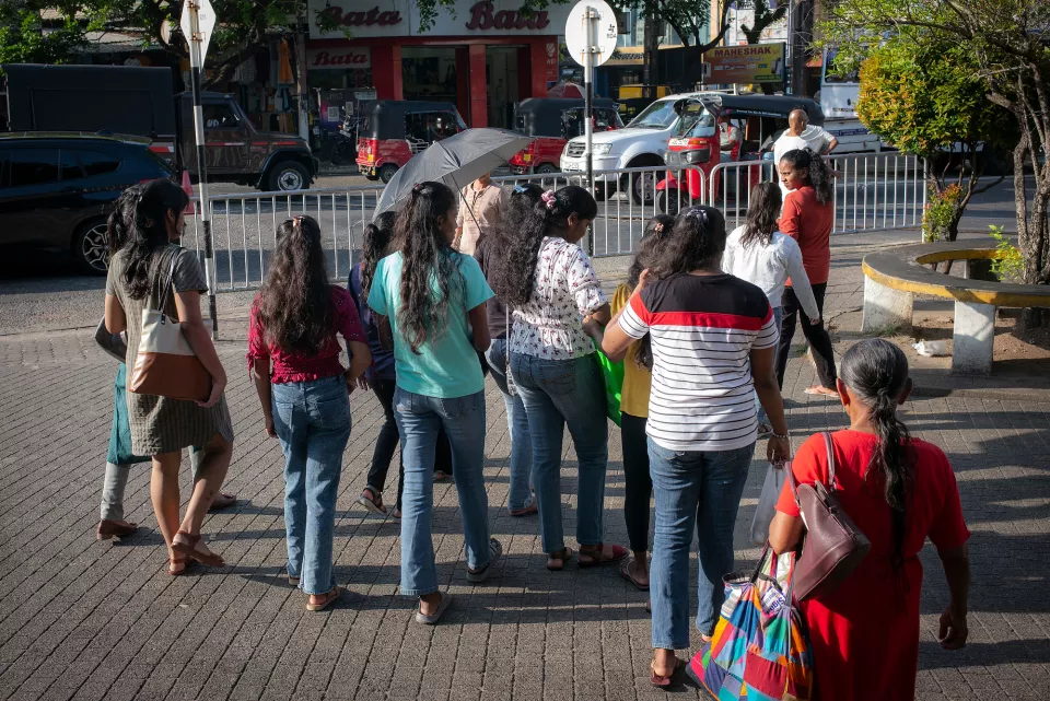 A group of women wait together at a pedestrian crossing in central Colombo, one shielding herself with an umbrella in the bright midday sun.