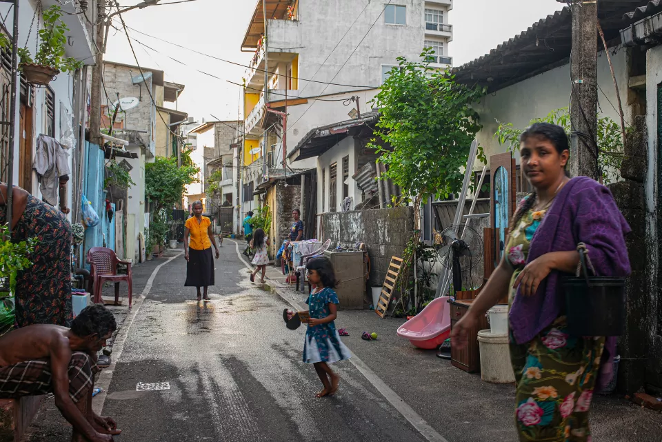 A shirtless man watches two young girls play in front of a vivid green and red street shrine, a Sri Lankan flag flying overhead.