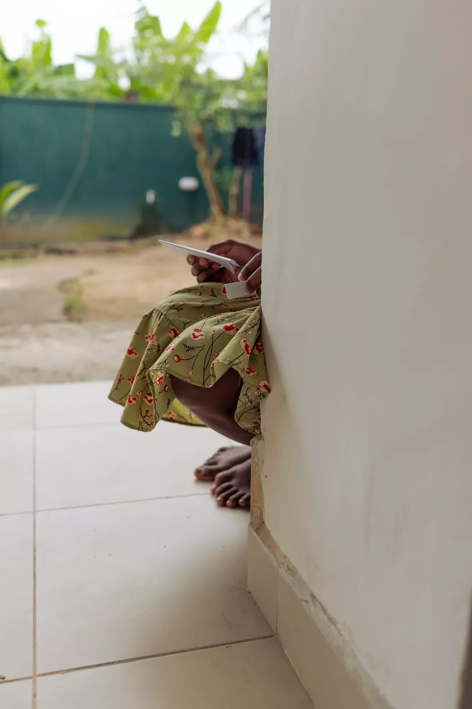 A person in a floral dress sits barefoot just around a doorway corner, quietly scrolling on a phone in a peaceful courtyard setting.