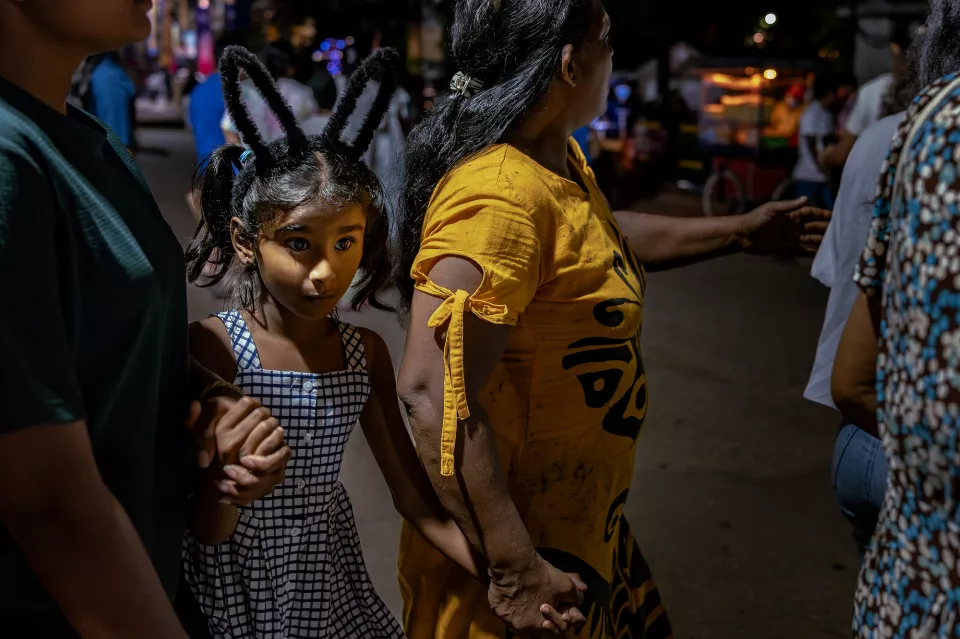 A young girl wearing bunny ears looks back with wide eyes while being guided through the busy night crowd by an adult.
