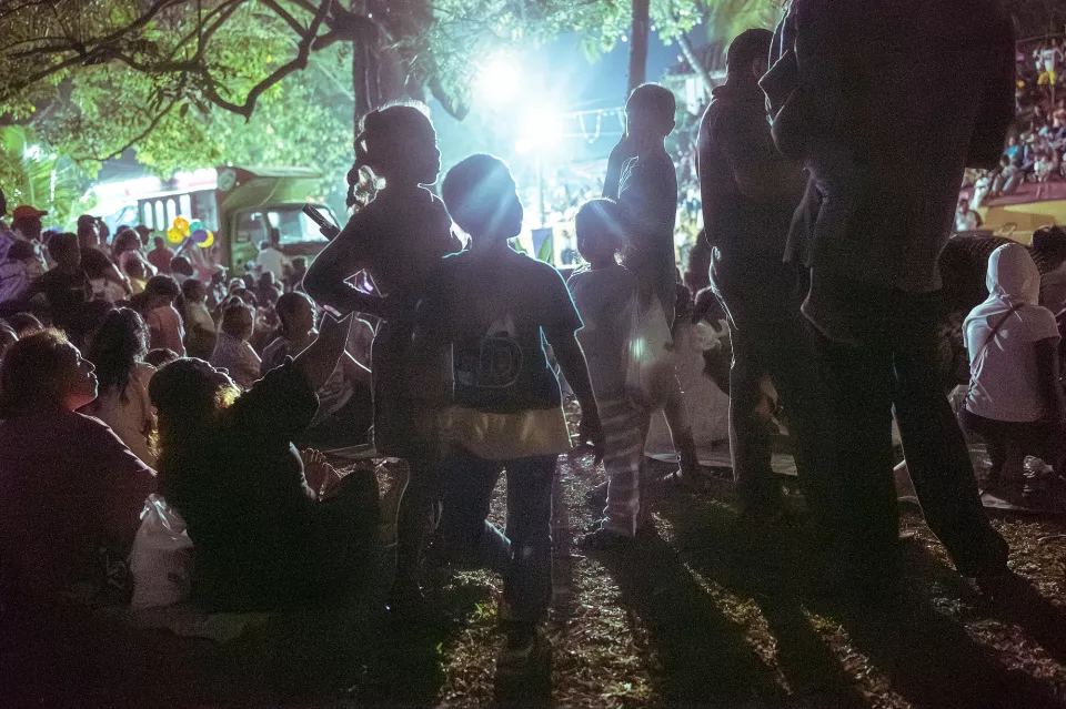 Children stand as dark silhouettes against a bright floodlight, surrounded by a large seated crowd at an outdoor night festival in Colombo.