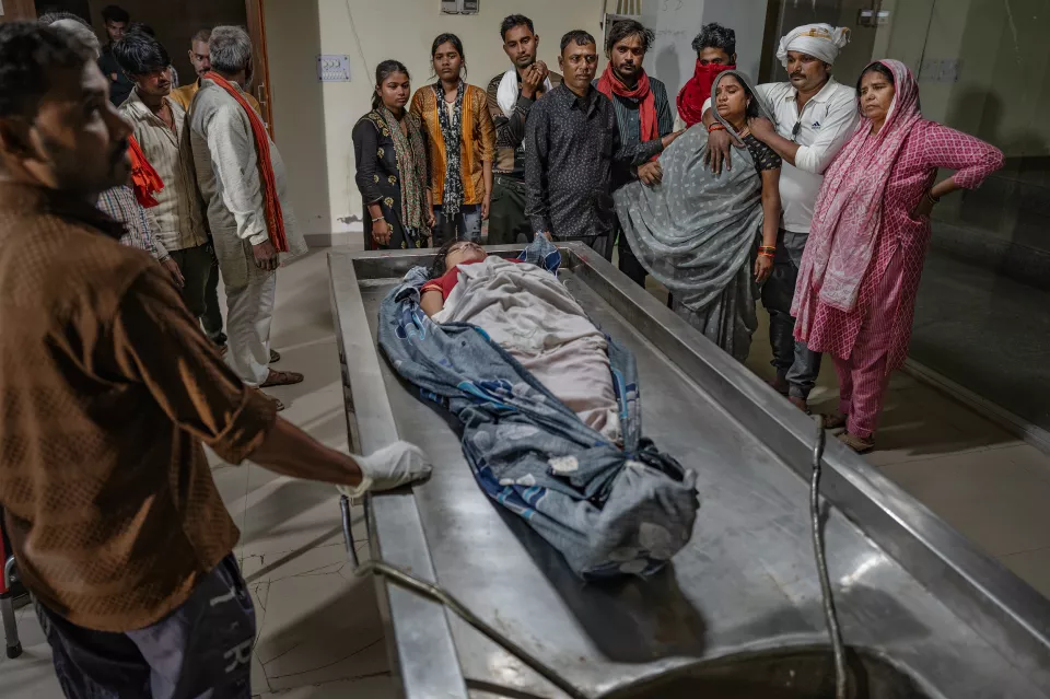Relatives gather around a body wrapped in cloth on a steel morgue table; a woman is held up by those beside her.