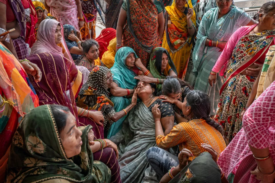 Women in colorful saris sit on the ground, consoling a grieving woman who is lying back in their arms.