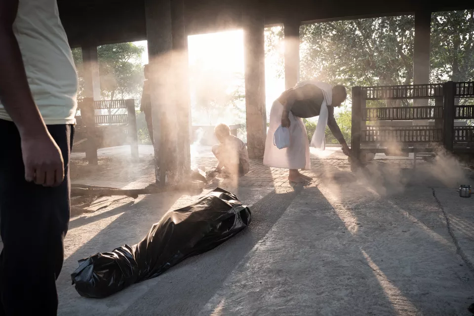 A body wrapped in black plastic lies on the ground of an open-air cremation shelter as a man sweeps in the smoky morning light.