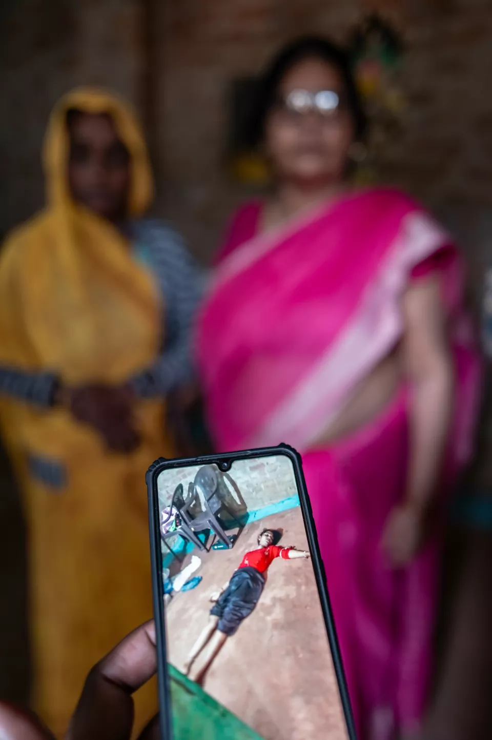 A hand holds a phone showing a photo of a young person lying on the floor, with two blurred women standing behind.