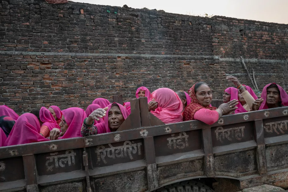 Women in pink saris ride in a tractor trailer in front of a brick wall, some looking toward the camera.