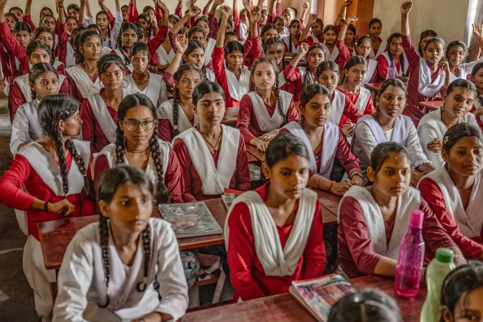Women in sheer pink saris stand close together in sunlight; one peers out from behind the fabric, and a girl stands nearby.