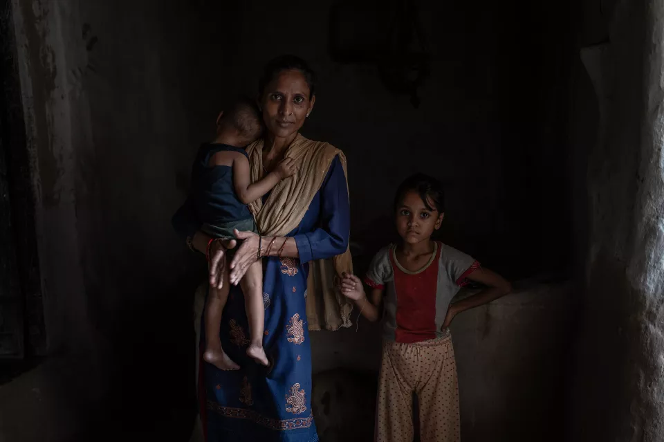 A woman holding a small child stands in a dark room beside an older girl who looks at the camera.