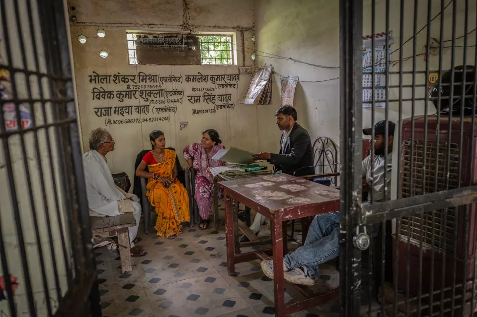 A man in a dark jacket reviews documents with two women and an older man in a small barred legal office.