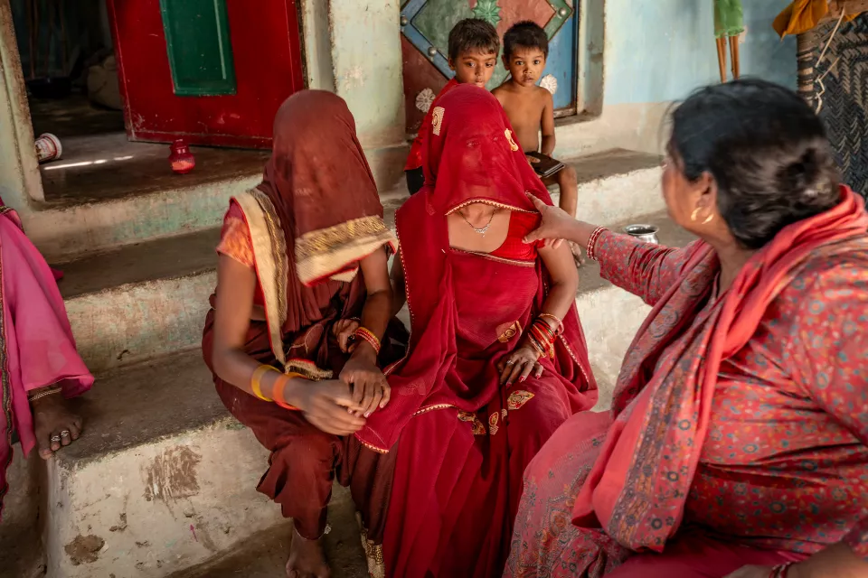 An older woman in red touches the arm of a veiled woman in a red sari seated on steps, with two boys watching behind.
