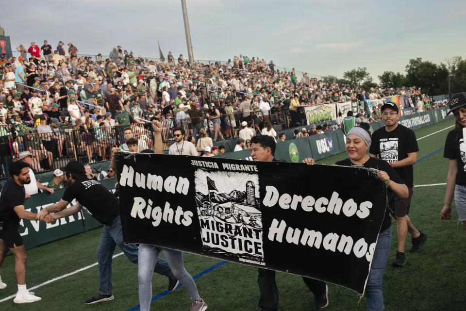 Two people wearing Migrant Justice t-shirts laughing together on the field, blurred crowd in the background.