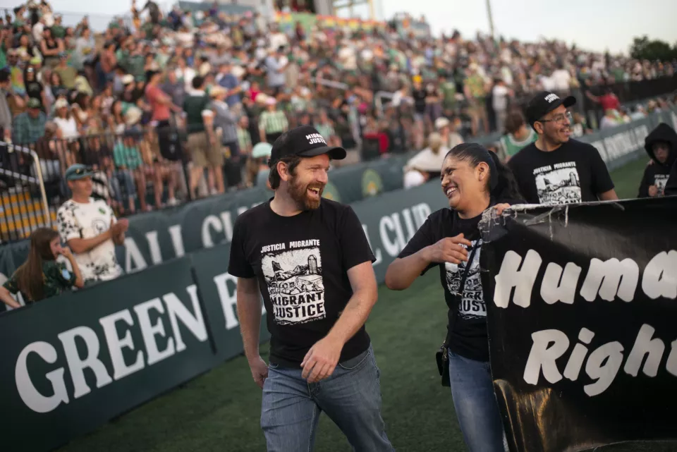 Soccer field at sunset; players on the pitch, spectators in the stands, and a group carrying a "Human Rights / Derechos Humanos &ndash; Migrant Justice" banner walks onto the field with a QR code sign.