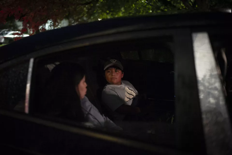 A woman in a car looks at a boy sitting in the back seat, nighttime scene, dark car window.
