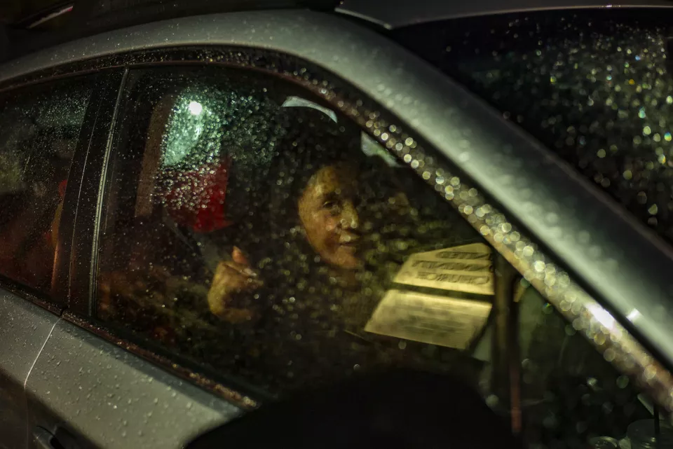 An elderly person's face reflected in a rain-streaked car window at night, "United Community Church" sign visible.