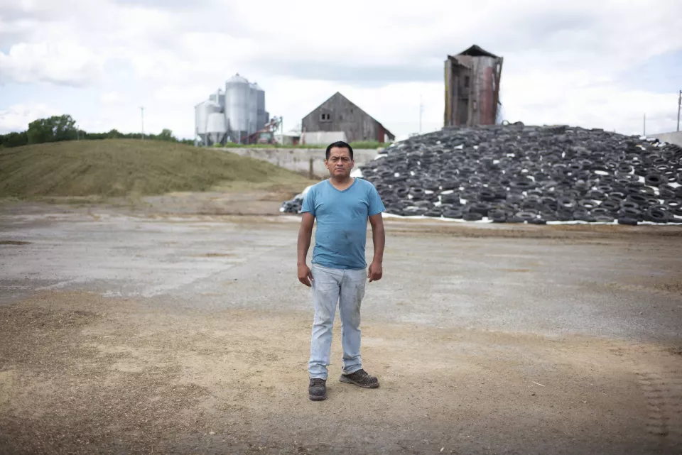 A man standing in a farmyard, pile of tires and silos in the background.