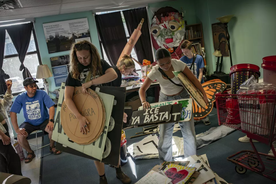 Two women pack up cardboard protest signs reading "ICE YA BASTA" and "&iexcl;NO M&Aacute;S... PO MIGRA" in a cluttered office, a man in a "Migrant Justice" t-shirt seated nearby, a butterfly costume prop in the background.