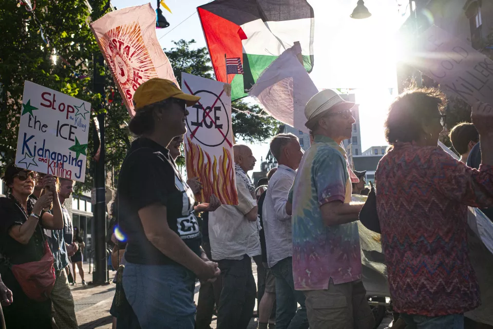 Crowd marching with signs reading "Stop the ICE Kidnapping and Killing" and a crossed-out "ICE" with flames, a Palestinian flag and American flag overhead, backlit by the sun.