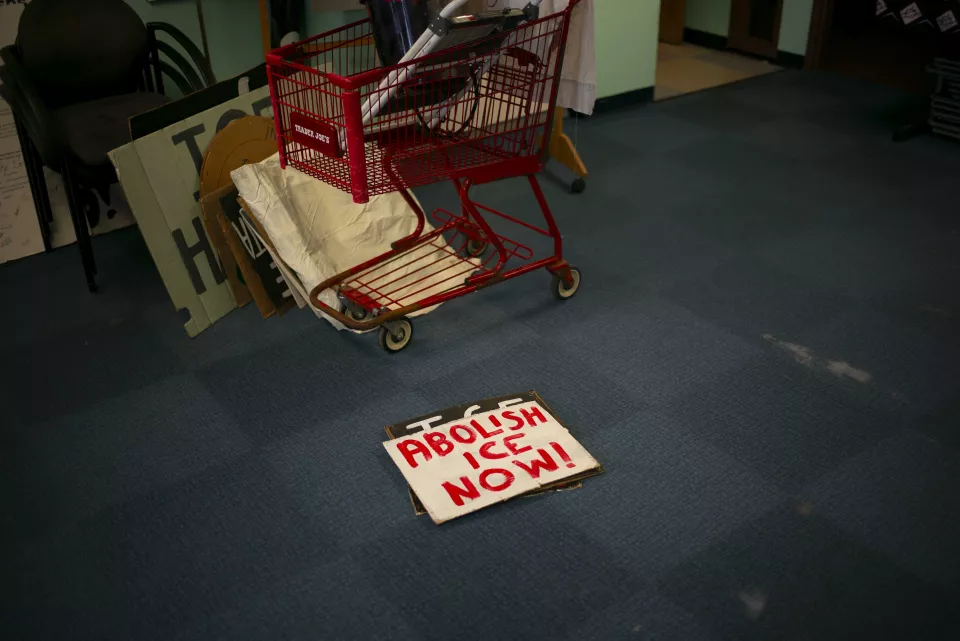 A red shopping cart loaded with protest signs sits in a dim office, a sign reading "ABOLISH ICE NOW!" lying on the floor nearby.