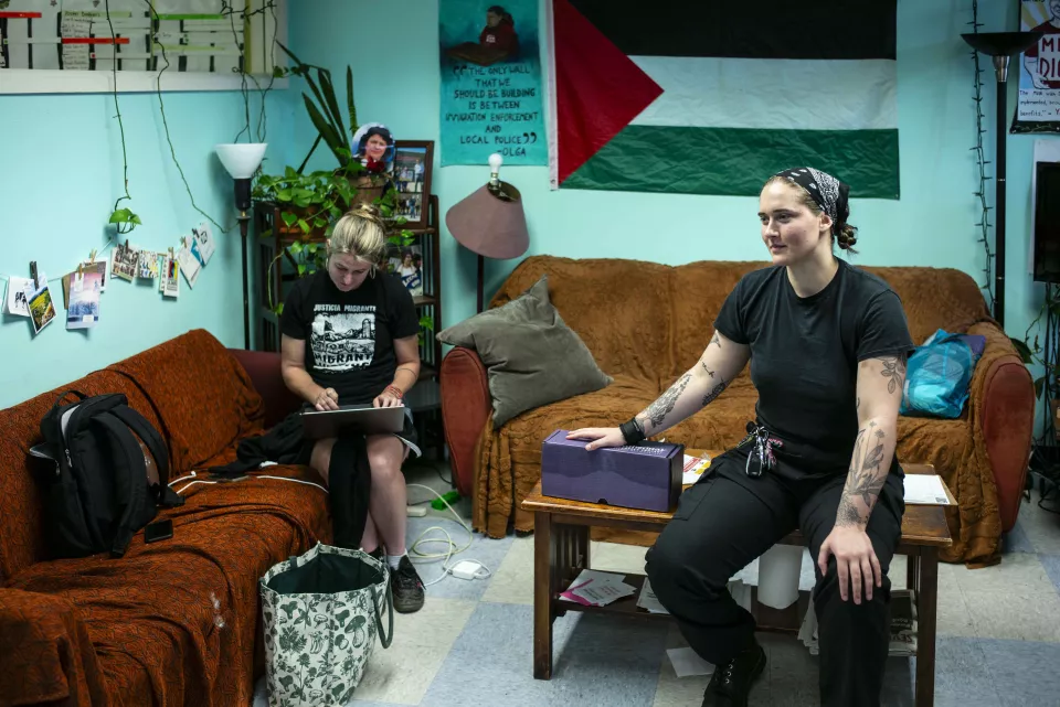 Two women in a living-room-style office space, one working on a laptop wearing a "Justicia Migrante" t-shirt, the other sitting with visible tattoos, a Palestinian flag on the wall behind them.