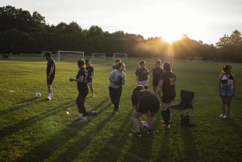 Family picnic in a park at sunset, people seated at a picnic table and in folding chairs near soccer goals, coolers and food spread on a table.