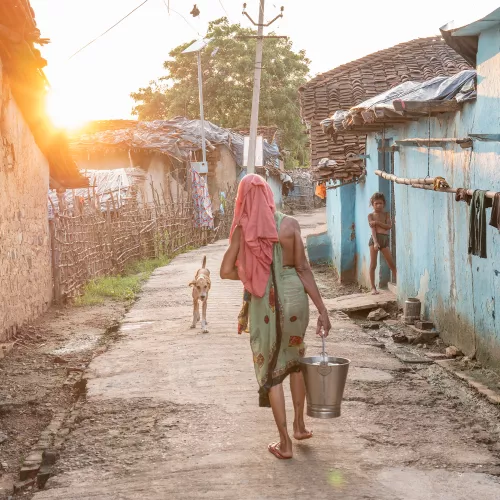 A woman carrying a metal bucket walks down a sunlit village lane toward a dog, while a child watches from a blue doorway.