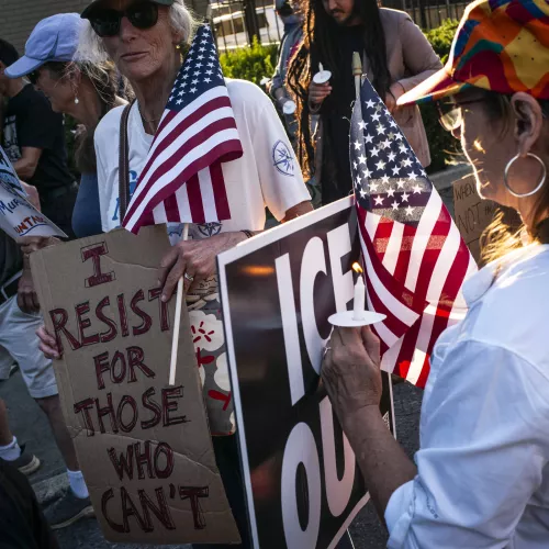Close-up of a woman lighting a candle for another woman holding a sign reading "I RESIST FOR THOSE WHO CAN'T," American flags and an "ICE OUT" sign nearby.