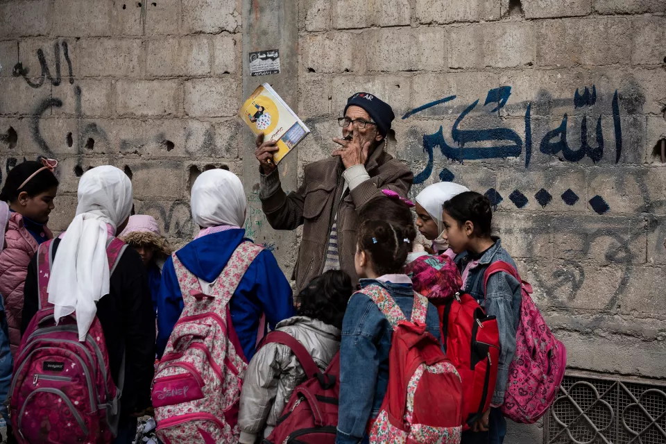An elderly man smoking a cigarette in front of a wall with blue Arabic writing shows a school notebook to young girls with colorful backpacks.