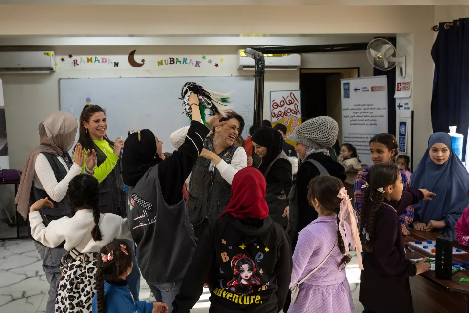 A tight-knit group of young girls, children, and volunteers, some veiled and some not, laugh together in a multipurpose room. 