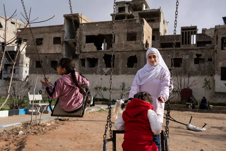 exterior of destroyed city - a woman pushes two young girls on a swing 