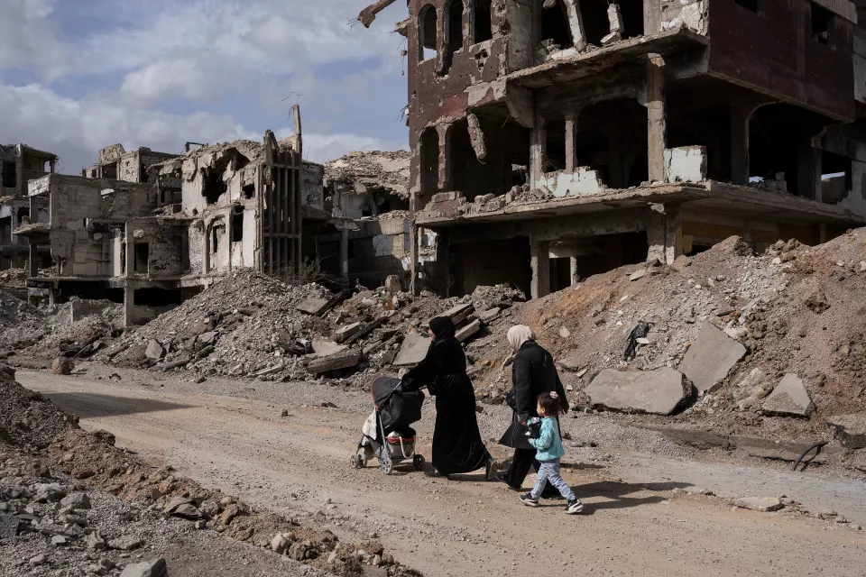 Outside scene of a destroyed city - two women dressed in black are pushing a stroller and walking briskly alongside a child dressed in light blue. 