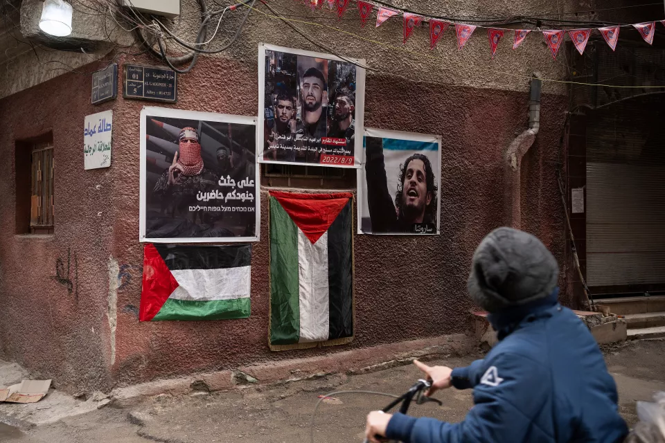 outside - in the foreground, a child on a bicycle looks at a wall displaying Palestinian flags and large photos of fighters 